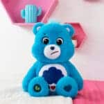 A blue Grumpy Bear plush toy from the Care Bears collection sits on a neatly made bed. The toy features a rain cloud with raindrops and hearts on its belly.