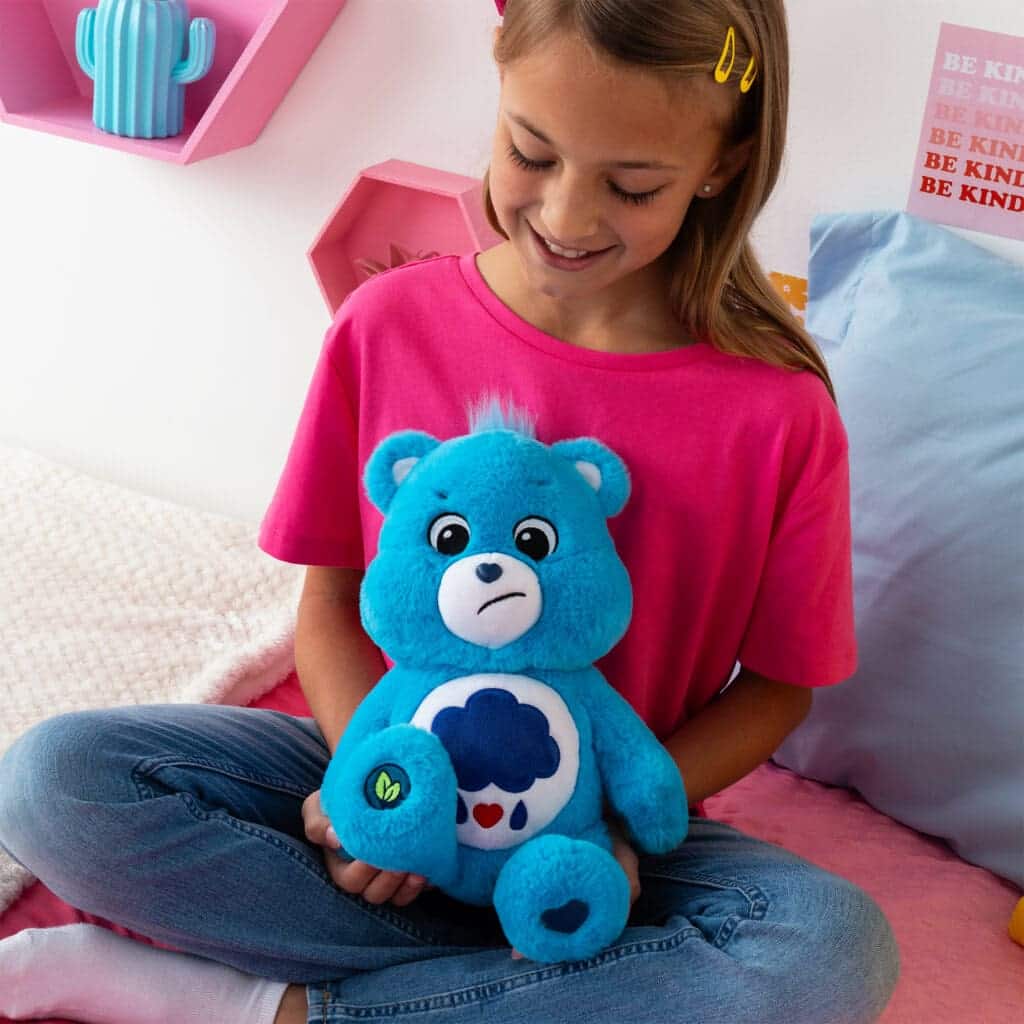 A young girl sits on a bed, smiling as she holds a blue Grumpy Bear plush toy from the Care Bears collection. The plush toy features a rain cloud with raindrops and hearts on its belly.