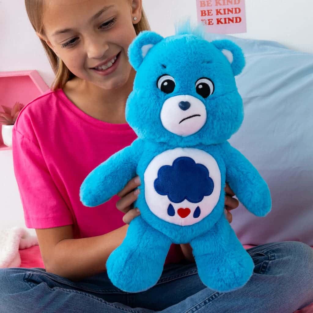 A young girl sits on a beige couch holding a Grumpy Bear plush toy from the Care Bears collection. The plush toy features Grumpy Bear's signature blue color and a rain cloud with raindrops and hearts on its belly, while the girl smiles warmly.