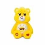A yellow Funshine Bear plush toy from the Care Bears collection is shown against a plain white background. The toy features a smiling sun on its belly and has a cheerful expression.