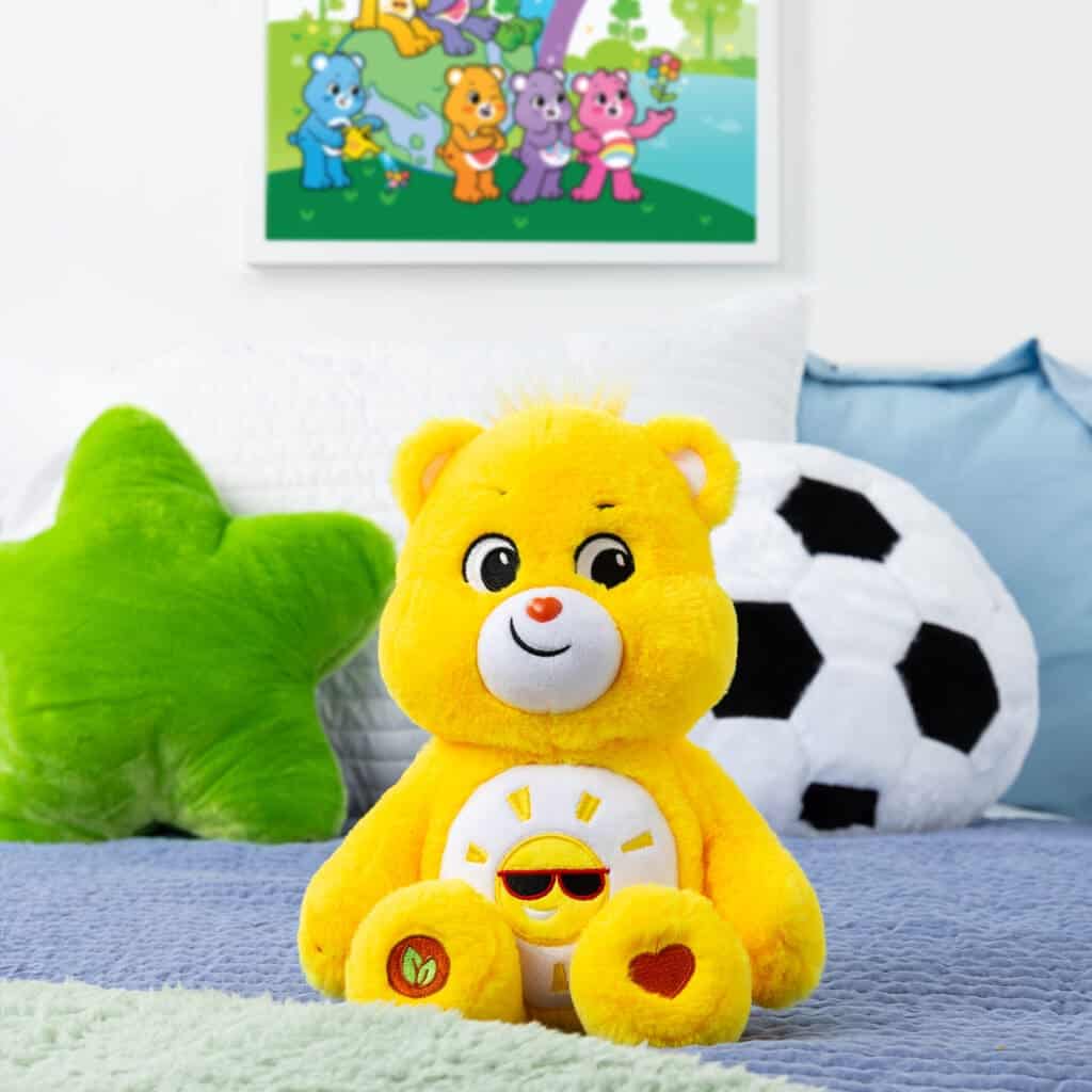 A yellow Funshine Bear plush toy from the Care Bears collection is placed on a bed. The plush toy features a smiling sun on its belly and has a cheerful expression.