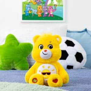 A yellow Funshine Bear plush toy from the Care Bears collection is placed on a bed. The plush toy features a smiling sun on its belly and has a cheerful expression.