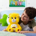 A yellow Funshine Bear plush toy from the Care Bears collection is placed on a bed with a young boy laying down. The plush toy features a smiling sun on its belly and has a cheerful expression.