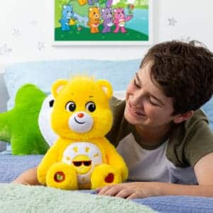 A yellow Funshine Bear plush toy from the Care Bears collection is placed on a bed with a young boy laying down. The plush toy features a smiling sun on its belly and has a cheerful expression.