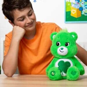 A green Good Luck Bear plush toy from the Care Bears collection is placed on a wooden table with a young boy. The plush toy features a four-leaf clover on its belly and has a cheerful expression, with a potted plant and a small decorative basket in the background.
