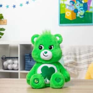 A green Good Luck Bear plush toy from the Care Bears collection is placed on a desk. The plush toy features a four-leaf clover on its belly and has a cheerful expression.