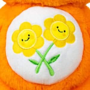 Close-up of an orange Friend Bear plush toy from the Care Bears collection, highlighting its embroidered two intertwined smiling flowers on its belly.