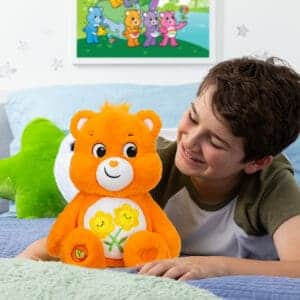 An orange Friend Bear plush toy from the Care Bears collection is placed on a white bed with a young boy. The plush toy features two intertwined smiling flowers on its belly and has a cheerful expression.