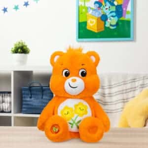 An orange Friend Bear plush toy from the Care Bears collection is placed on a table. The plush toy features two intertwined smiling flowers on its belly and has a cheerful expression.