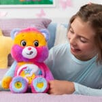A young girl sits on a white bed, smiling and holding a purple Dare to Care Bear plush toy from the Care Bears collection.
