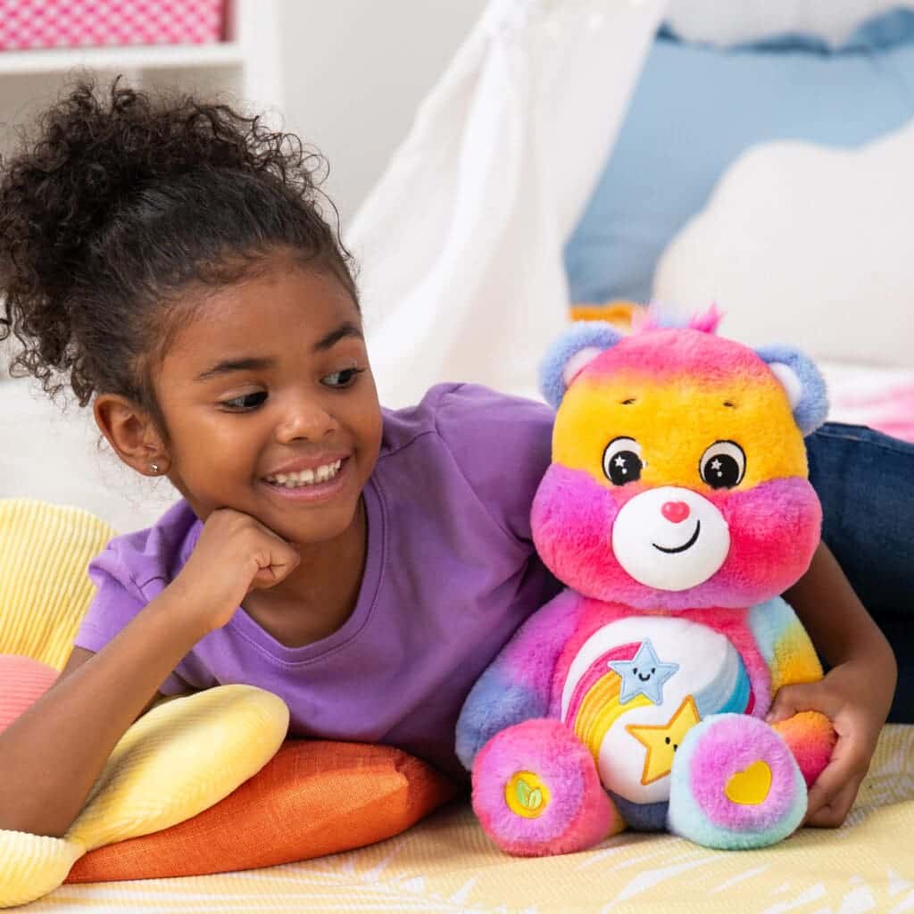 A Dare to Care Bear plush toy from the Care Bears collection is placed on a bed with a young girl smiling.