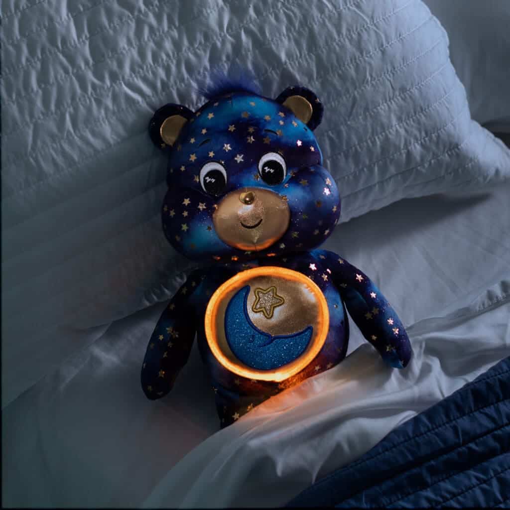 Care Bears Collector Edition Bedtime Bear has a dark blue star filled body with a gold moon stomach emblem laying on bed with white sheets.