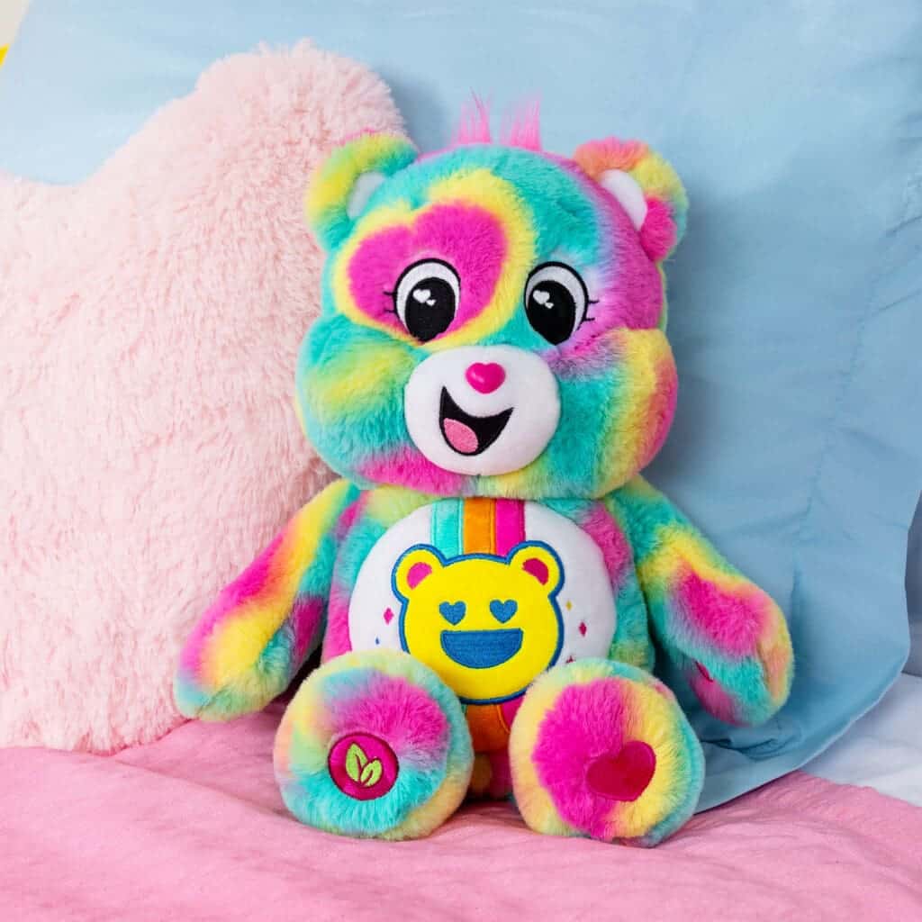 A colorful Good Vibes Bear plush toy from the Care Bears collection sitting on a bed.