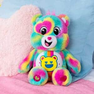 A colorful Good Vibes Bear plush toy from the Care Bears collection sitting on a bed.