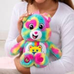 A colorful Good Vibes Bear plush toy from the Care Bears collection is being held by a young girl.