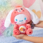 Hands holding Care Bears Funsize Cinnamoroll plush displayed in a lifestyle setting, featuring a cute and cuddly design with a pink heart emblem on its stomach, promoting love and friendship.
