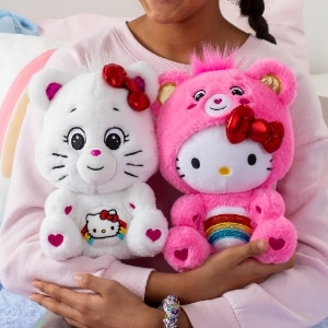 Care Bears Hello Kitty white and pink plush toys with rainbows on their stomachs, held by a child's hand.